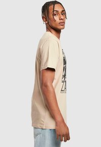 Beige bavlněné tričko s krátkým rukávem, které má grafický potisk na přední straně. Design zahrnuje černý text a obrázky.