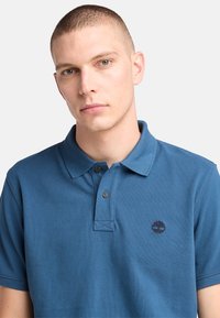 Blå pikétröja i texturerad bomull, med en plagg inlägg med två knappar och en liten broderad logotyp på bröstet. Korta ärmar.