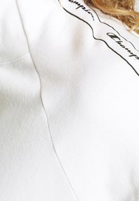 Felpa bianca caratterizzata da un tessuto morbido e testurizzato, con un logo nero stampato sul petto. Le cuciture visibili aggiungono un tocco di dettaglio.