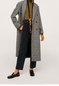 Abrigo gris con un diseño de doble botonadura, cuello de tortuga oliva, jeans oscuros y mocasines burdeos. Mangas largas y corte relajado.