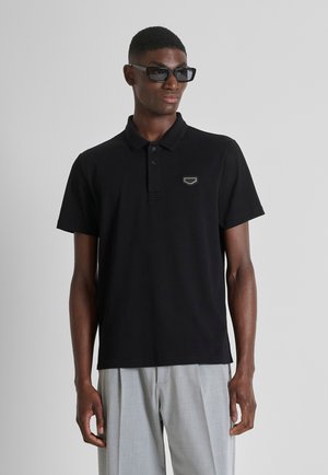 Homme portant un polo noir à manches courtes avec un patch logo, un pantalon gris plissé et des lunettes de soleil rectangulaires noires, debout devant un fond uni.