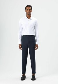 Weißes Langarmhemd mit Knöpfen, kombiniert mit dunkelblauen Hosen mit elastischem Bund. Schwarze Loafer vervollständigen das Outfit.