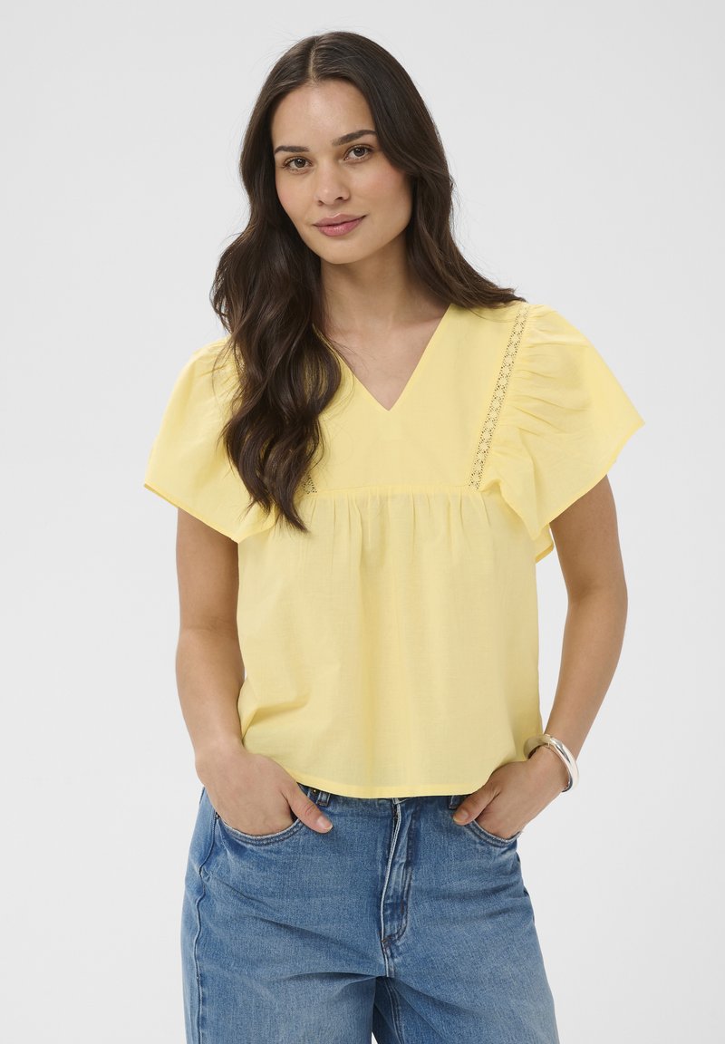 Femme aux longs cheveux foncés portant un chemisier jaune à col en V avec des manches à volants et un jean bleu, mains dans les poches devant un fond uni.