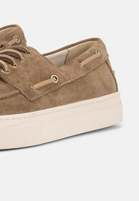 Καφέ σουέντ casual παπούτσια σκάφους με μπεζ κορδόνια, μεταλλικές τρύπες και λευκές λαστιχένιες σόλες σε λευκό φόντο.