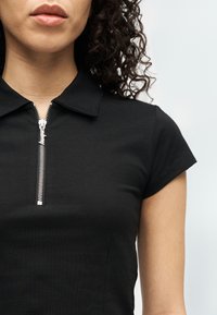 Γυναίκα με σγουρά μαλλιά που φοράει μαύρο τοπ με κοντά μανίκια, ασημί φερμουάρ και γιακά, μπροστά σε απλό φωτεινό φόντο.