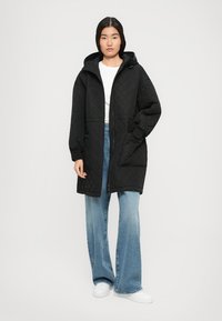 Vrouw in een zwarte gequiltde hooded jas, lichtblauwe wijde broek, witte sneakers en een wit T-shirt, staande tegen een witte achtergrond.