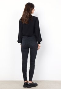 Svart långärmad blus med puffärmar, matchad med smala svarta jeans. Modellen bär kraftiga svarta skor. Enfärgad bakgrund.