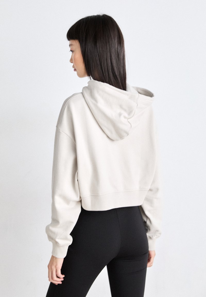 Beige P cropped Hoodie aus weichem Material, mit Kapuze und Kordelzug sowie gerippten Bündchen, kombiniert mit schwarzen, enganliegenden Leggings.