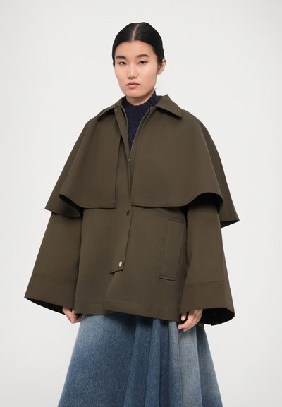 Olive green elongated jacket with a cape-like overlay, featuring a button closure and large front pockets. Blue denim-like layered skirt.