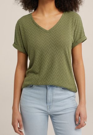 Vrouw die een olijfgroene, gestructureerde V-hals top met korte mouwen draagt, ingestopt in lichtblauwe spijkerbroek, staand met ontspannen armen langs het lichaam.