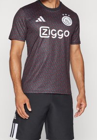 Černý a červený vzorovaný sportovní dres s kulatým výstřihem a krátkými rukávy, s bílým logem a textem na přední straně, vyrobený z lehkého materiálu.