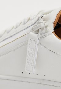 Hvide lædersneakers med en polstret krave, snørebånd og et mærke-tag på siden, der viser minimalistisk stil og håndværk.