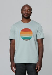 T-shirt bleu clair avec un graphique circulaire comportant des bandes horizontales dans des tons d'orange, rouge et bleu-vert. Manches courtes et design à col rond.