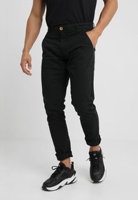 Hombre vistiendo pantalones negros ajustados con los puños arremangados, una mano en el bolsillo, combinados con zapatillas negras y blancas sobre un fondo liso.