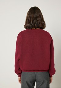 Vyno raudonos spalvos puloveris su ilgomis rankovėmis, tekstūruota medžiaga, susigūžęs kaklas ir apatinė briauna, derinamas su pilkais kelnais. Pavaizduota vaizdas iš nugaros.