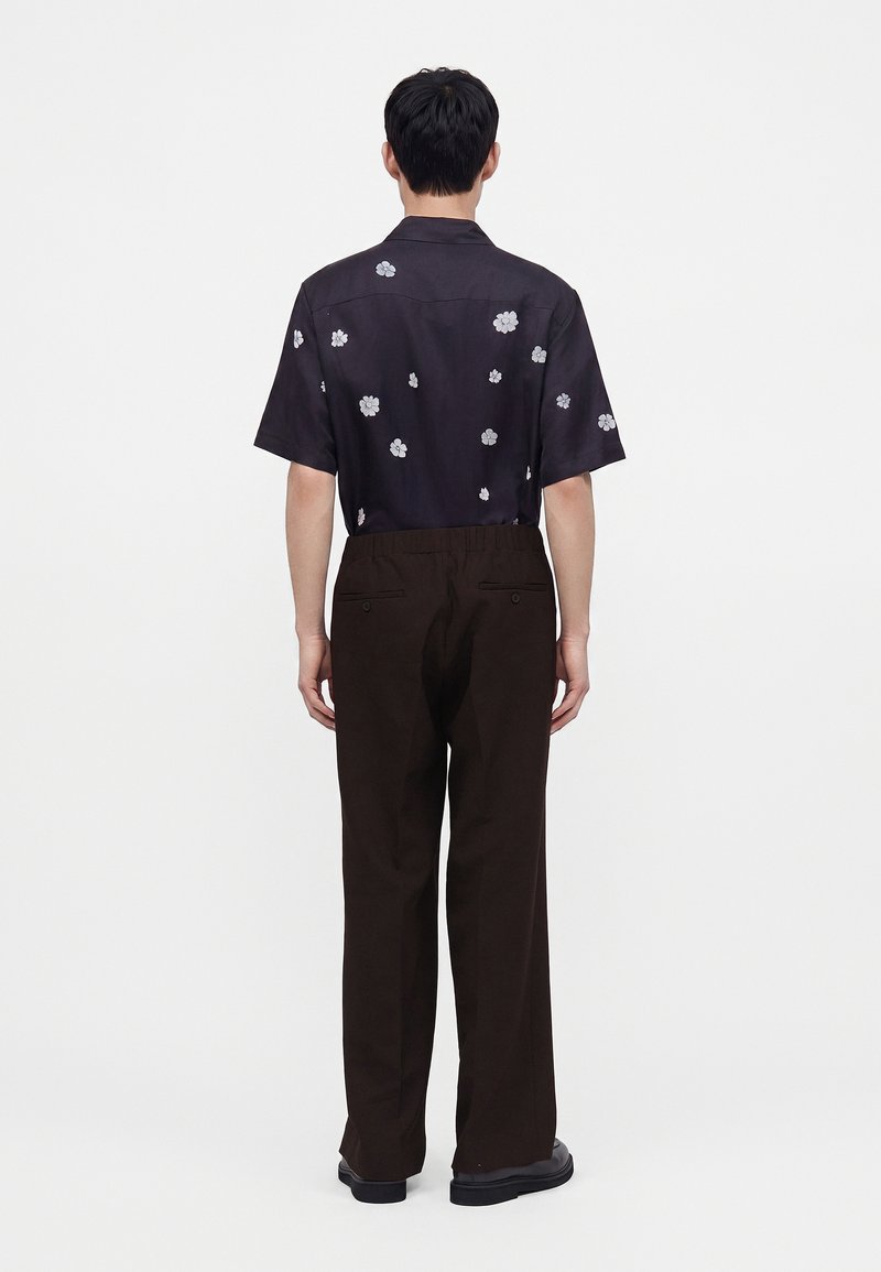 Hombre que lleva una camisa negra de manga corta con estampados de flores blancas y pantalones sueltos marrones oscuros, de pie de espaldas sobre un fondo blanco.
