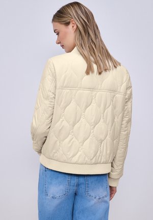 Kobieta o blond włosach do ramion, ubrana w kremową pikowaną kurtkę i niebieskie dżinsy, widziana od tyłu, zwrócona w lewo.