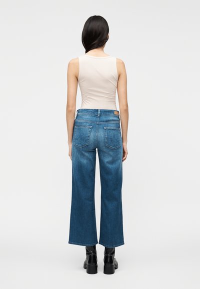 Canotta beige chiaro abbinata a jeans blu a gamba larga. Il denim ha un effetto sbiadito e presenta tasche posteriori e una toppa in pelle sulla vita.