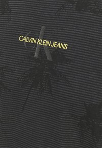 Černé bavlněné tričko s horizontálními pruhy, vzorem palmových stromů a žlutým logem "CALVIN KLEIN JEANS" s šedým akcentem "K".