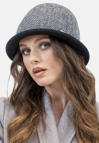 Chapeau cloche tissé noir et blanc avec une couronne arrondie et un large bord. Texture douce ; associé à un manteau gris clair.