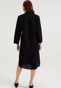 Černé lněné šaty s límečkem, dlouhými rukávy a rovnou siluetou. Spodní lem je mírně zakřivený, odhalující detail dosahující k kotníkům.