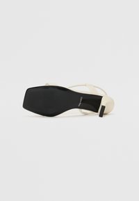 Mustassa ja kermaisessa slingback-sandaalissa on neliön muotoinen varvasosa, litteä korko ja kapeat hihnat. Pintamateriaali on sileää synteettistä materiaalia, jossa on teksturoitu pohja.