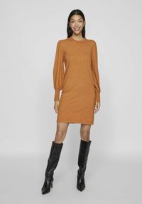Orange stickad klänning med långa, puffiga ärmar och rundad halsringning. Ett par svarta knähöga klackstövlar kompletterar outfiten.