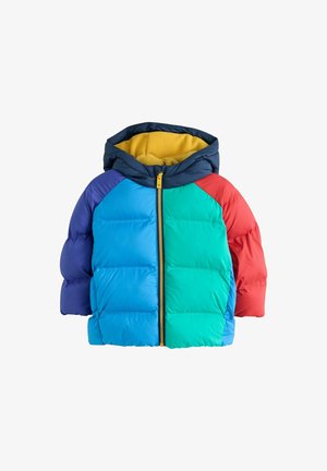 Ung dreng iført en farverig dynejakke med blå, grønne, røde og lilla sektioner, blå jeans og hvide sneakers, står og kigger mod højre.