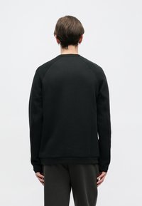 Joven con cabello corto castaño, vistiendo una camiseta de manga larga negra y pantalones oscuros, de espaldas contra un fondo blanco liso.
