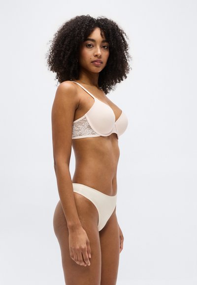 Γυναίκα με σγουρά μαλλιά που φοράει γαλλικό σουτιέν με δαντελένια πλάτη σε γυμνό χρώμα και αντίστοιχο εσώρουχο, στέκεται σε πλευρικό προφίλ μπροστά σε απλό λευκό φόντο.