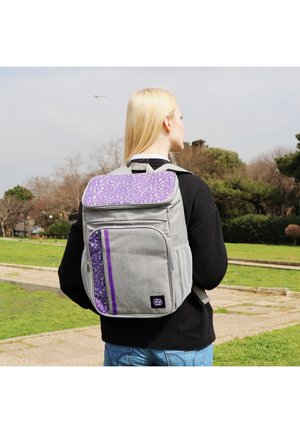 Person with blonde hair wearing black hoodie and jeans, carrying a grey and purple backpack, standing on a park path with trees in background.