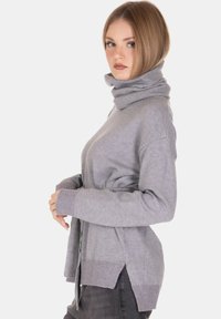 Giovane donna con i capelli biondi corti, che indossa un maglione grigio a collo alto e pantaloni scuri, in piedi di profilo su uno sfondo bianco.