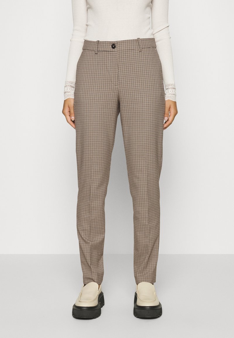 Vrouw in beige en zwart geblokte tailored broek met een knoop in de taille, gecombineerd met crèmekleurige instappers en een witte aansluitende trui.