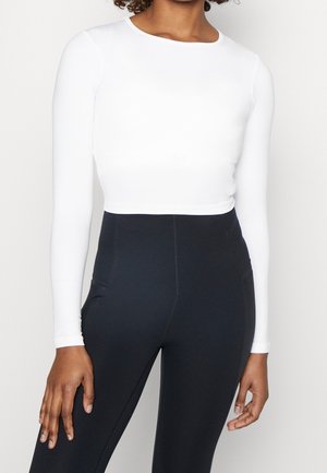 Kobieta ubrana w dopasowaną, białą, długorękawową krótką bluzkę i czarne legginsy z wysokim stanem, stojąca na tle jednolitego tła.