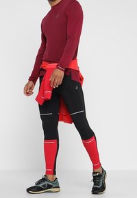 Bordowy długi rękaw sportowy top z fakturowanej tkaniny, w połączeniu z czarnymi i czerwonymi legginsami z odblaskowymi akcentami. Czarne buty sportowe.