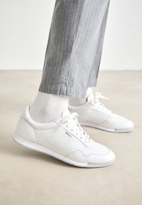 Hvite lær sneakers med teksturerte detaljer, en logo på siden, og gummisåle. Kombinert med grå stripete bukser og hvite sokker.