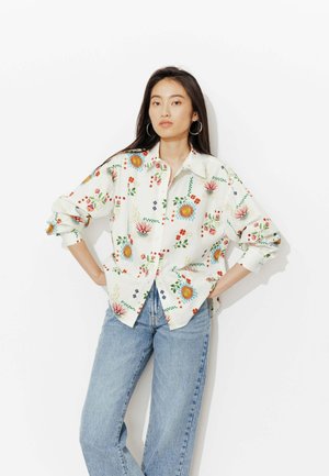 Femme portant une chemise blanche oversize à boutons avec des imprimés floraux colorés et un jean bleu clair, debout les mains sur les hanches devant un fond blanc.