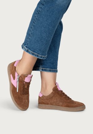 Beine in blauen kurzen Jeans und braunen Wildledersneakern mit rosa Akzenten und seitlichem Reißverschluss, stehend auf neutralem Hintergrund.