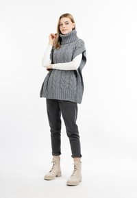 Grauer gestrickter Poncho mit hohem geripptem Kragen und einem Zopfmuster. Über einem weißen Langarmoberteil getragen, kombiniert mit dunklen Hosen und beigen Stiefeln.
