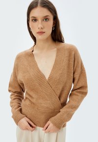 Ruskea, kevyt neulepusero, jossa on kietaisusuunnittelu, syvä V-neck ja ribbaus hihansuissa ja helmassa. Pehmeä rakenne, rento istuvuus.