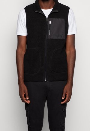 Man i svart zip-up fleeceväst med bröstficka över vit kortärmad skjorta och svarta cargobyxor mot en enfärgad bakgrund.