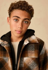 Brown and white plaid jacket with a textured feel, featuring a black hooded inner layer and a zippered front closure.