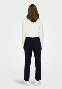 Námořnické kalhoty na míru s slim fit, dvěma zadními kapsami a čistým pasem, v kombinaci s bílým tričkem s dlouhým rukávem a bílými teniskami.