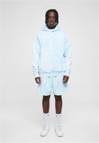 Svetlomodrý hoodie s tie-dye vzorom a predným vreckom, v kombinácii s rovnocennými šortkami. Bavlnená látka, uvoľnený strih, rebrované manžety a biele tenisky.