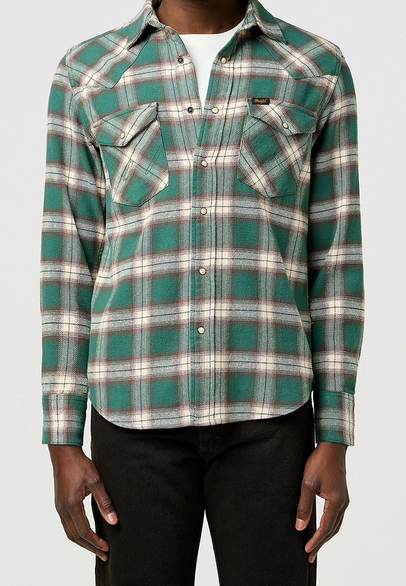 Mand iført grøn og hvid ternet skjorte med brystlommer over en hvid undertrøje, kombineret med sorte bukser.