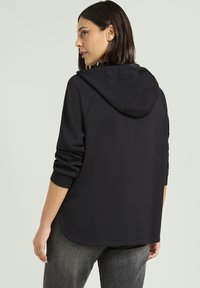 Felpa nera con una vestibilità rilassata, maniche a raglan e un ampio cappuccio. Texture morbida e orlo curvo, indossata con jeans grigi.