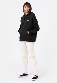 Black hooded sweatshirt with a front pocket and logo detail, paired with light beige pants and black sneakers. Smooth fabric texture.