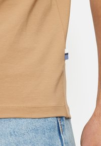Beige Baumwoll-T-Shirt mit tailliertem Schnitt, das an der Seitennaht ein kleines blau-weißes Akzentetikett und eine glatte Textur aufweist.