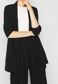 Blazer noir ouvert avec une coupe décontractée, une texture douce et un devant légèrement drapé. Porté sur un haut blanc, associé à un pantalon noir.