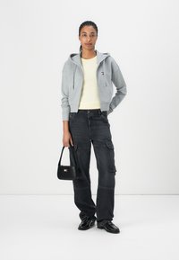 Felpa grigia corta, camicia gialla, pantaloni cargo neri e scarpe nere. Piccola borsa con logo. Sfondo bianco semplice.